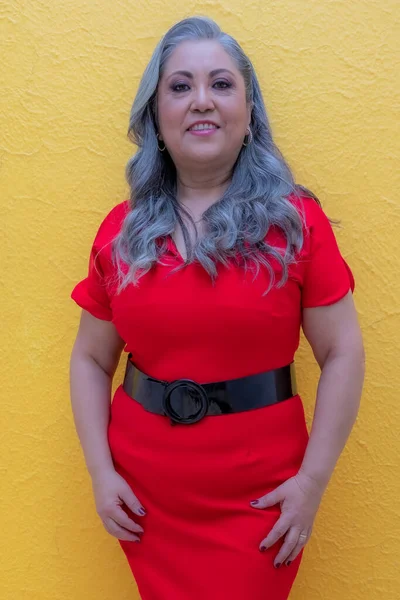 Frontal portrait of a smiling woman standing against a yellow background, long gray hair, red dress with a black belt, modern make-up in warm colors