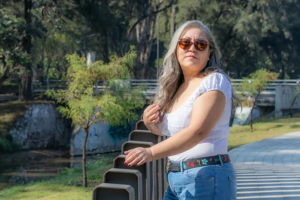 Smiling senior adult woman standing enjoying sunny day, bridge, stream and trees with green foliage in blurred background, long gray hair, sunglasses, white blouse, jean, sunny day in the park