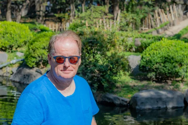 Portrait of handsome dutch senior adult man wearing sunglasses and blue blouse, pond, green bushes and trees in blurred background, enjoying sunny day in Japanese garden, looking at camera