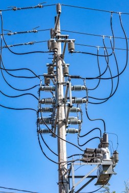 Elektrik kabloları, kondüktörleri, vanaları ve yalıtıcılarıyla yüksek voltaj direği arka planda mavi bir gökyüzüne karşı, güneşli bir günde düşük perspektiften görülüyor.