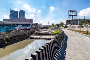 Zapopan, Jalisco Meksika. 1 Ocak 2023. Açık kanalizasyon kanalı boyunca boş yaya geçidi ve bir araç köprüsü, arka planda mavi gökyüzüne karşı modern binalar. Güneşli bir günde şehir manzarası