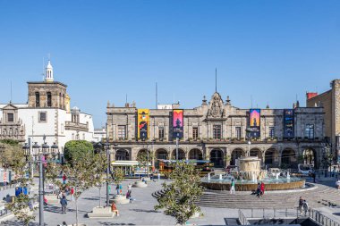 Guadalajara, Jalisco Meksika. 4 Şubat 2023. Plaza de los Laureles şehrinin manzarasında insanlar yürüyor, tur otobüsleri, ağaçlar, bir çeşme ve arka planda Palacio belediye binası güneşli bir günde mavi gökyüzü