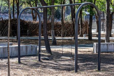 Kemerli metal tüp tabanında iki salıncak, halka açık bir parkta çocuklar için açık alan, bulanık arka planda yeşil yapraklı ağaçlar, Meksika 'da güneşli bir gün.