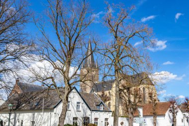 Beyaz duvarlı çıplak ağaçlar ve arka planında mavi gökyüzü ve beyaz bulutlu çan kulesi olan St. Michael Manastırı kilisesi Midden-Limburg, Hollanda 'da güneşli bir gün.