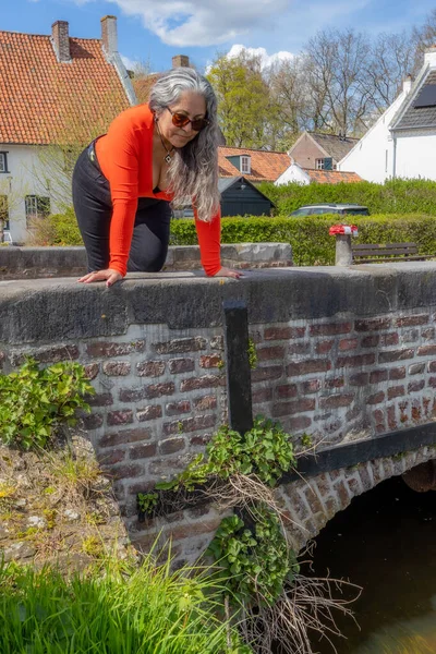 Latin American woman crouching and leaning over a bridge looking down, houses with white walls in background, long wavy gray hair, orange low-cut top and black pants, sunglasses, Thorn, Netherlands