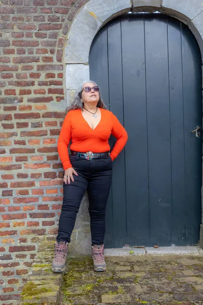 Latin American senior adult woman standing and leaning against brick wall next to closed wooden door, looking up, long wavy gray hair, wearing low cut orange top, black pants and sunglasses