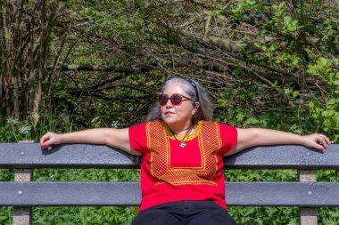 Latin esmer kadın, ahşap bir bankta rahat bir şekilde oturuyor, bulanık arka planda yabani yeşil bitki örtüsü, yerli sarı bluz, güneş gözlüğü, gri saçlar, güneşli bir günün tadını çıkarıyor, Hollanda