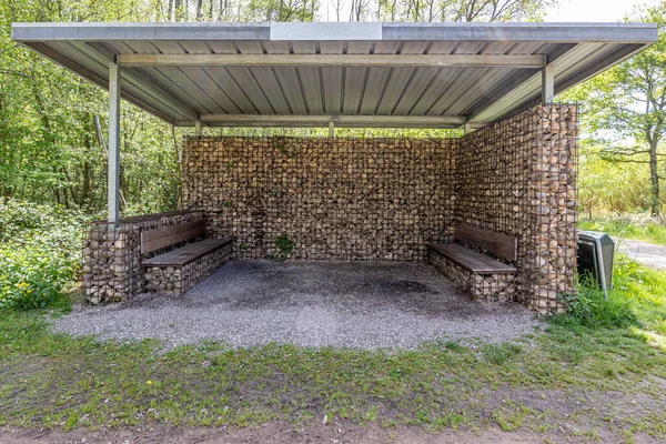 stock image Ecological gabion wall shelter, sheet metal roof and wooden benches, sunny day in IJzerenbos nature reserve, South Limburg, Netherlands. New technique in manufacture of retaining or decorative walls