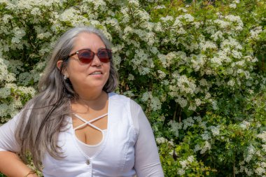 Uzun gri saçlı, sırıtan Latin Amerikalı esmer bir kadının belden yukarı portresi. Güneş gözlüğü takıyor, arka planda beyaz çiçekli bir bitki, beyaz günlük kıyafetler giyiyor, güneşli bir bahar gününün tadını çıkarıyor.