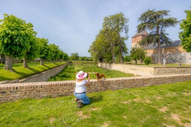 Şapkalı bir kadın cep telefonuyla Borgharen kale bahçelerindeki tuğla çitlerin üzerinde duran kısa saçlı daksunduna fotoğraf çekiyor yapraklı ağaçlar ve kuru hendek, Güney Limburg, Hollanda 'da güneşli bir gün.