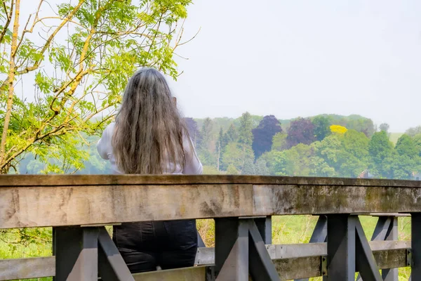 Olgun, gri saçlı bir kadın arkası kamerayla ahşap bir köprüde, sisli arka planda ağaçlar, beyaz bluz, güneşli bir bahar günü Ingendael, Güney Limburg, Hollanda 'daki doğa parkında duruyor.