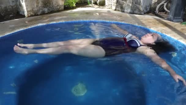 在墨西哥 一位年长的墨西哥妇女平静地漂浮在一个小池子里 挥动着胳膊 穿着全套游泳衣 身后有岩石墙的室外游泳池 阳光灿烂 — 图库视频影像