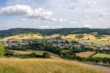 Almanya 'nın Rijnland-Palts eyaletinde yer alan Eiffel Kreis, Bitburg-Prum bölgesinde, tepeyi kaplayan yeşil yapraklı ağaçlar arasında küçük bir köy ile Alman kırsalının havadan görünüşü