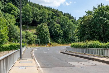 Arka planda trafik işaretleri olan boş bir köy yolu Stausee Bitburg nehri üzerindeki Staumauer Bitburger barajı üzerindeki araç köprüsü tepe üzerindeki yapraklı ağaçlar, Almanya 'da güneşli bir bahar günü.
