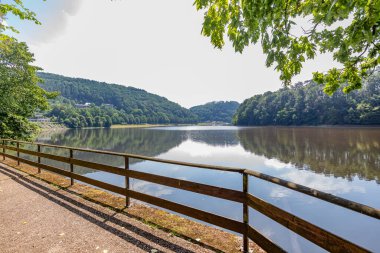 Stausee Bitburg Gölü 'nün etrafındaki yürüyüş parkurları, turist gözcülüğünde ahşap çitler, sisli arka planda bol yapraklı ağaçlarla dolu dağlar, Almanya' da mavi gökyüzü olan güneşli bir yaz günü.