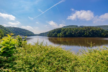 Stausee Bitburg gölünün manzarası turist gözcüleri, yeşil yabani bitkiler, arka planda bol yapraklı dağlar, güneşli bir yaz günü mavi gökyüzü ve beyaz bulutlar Almanya 'da