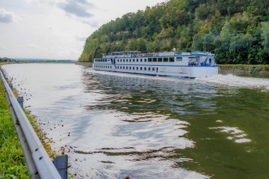 Albert Kanalı 'nın sularında yol alan turist teknesi, yeşil yapraklı ağaçlarla kaplı dağlar, arka planda ufuk, su yüzeyinde yansıma, Kanne, Riemst, Belçika' da gri bulutlu bir yaz günü.