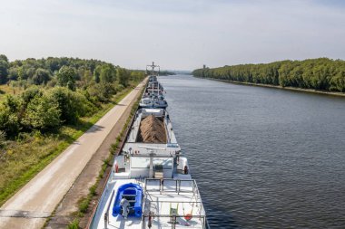 Albert Kanalı 'nın kıyısına demirlemiş kargo botları Lanaye Kilidi' ne, paralel boş kırsal sokağa, bol yapraklı yeşil ağaçlara, güneşli bir güne ve arka planda sisli mavi gökyüzüne ulaşmadan önce,