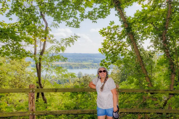 Mutlu gülümseyen kadın turist Caestert Platosu 'ndaki yemyeşil ağaçlar, Maas Nehri ve arka planda Hollanda kırsalları arasında uzanan uzun örgülü saçlar, Riemst, Belçika' da güneşli bir gün.