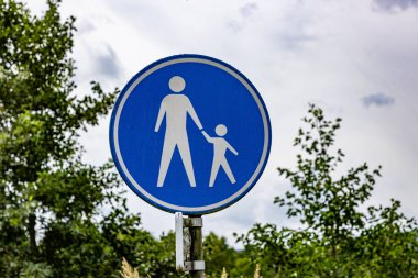 Yaya yolu veya yürüyüş alanı işareti bulutlarla kaplı gökyüzü ve arka planda yeşil yapraklar, mavi metalik yuvarlak kenarlık ve Hollanda 'da beyaz bir çocukla birlikte yürüyen bir yetişkinin çizimi