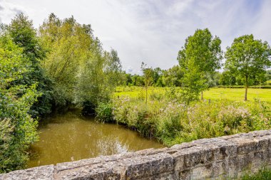 Geul Nehri, taştan köprüden görünen yabani bitkiler ve yapraklı ağaçların arasında, arka planda gri bulutlarla kaplı gökyüzüne karşı ovalar, Terpoorterweg, Epen, Güney Limburg, Hollanda 'da bulutlu bir gün