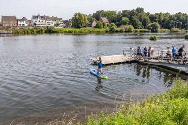 Lanaye, Vise, Belçika. 3 Eylül 2023. Maas Nehri 'nde sörf tahtasında genç bir adam, feribot rampasında bisikletleri olan insanlar, Hollanda köyü ve arka planda yapraklı ağaçlar, güneşli yaz günü