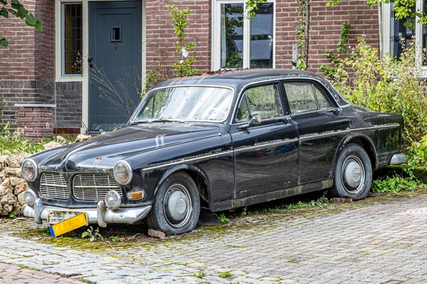 Old, run down, dirty and abandoned black car with flat tires, parked in front of a house with brick walls in background, surrounded by green wild vegetation