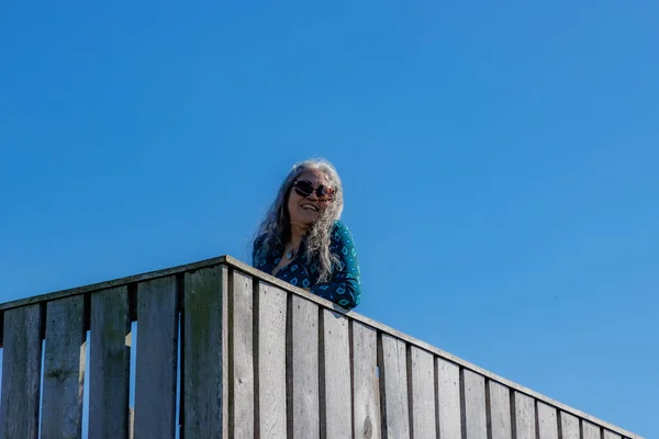 Smiling senior adult tourist leaning on wooden fence of observation tower, sunglasses, blue-green blouse, clear sky in background, sunny summer day