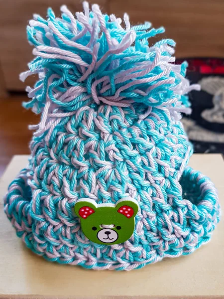 Small blue white hat with pompom for dog or cat crocheted on a wooden surface, green bear face button as decoration, blurred background. Handmade creativity concept