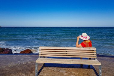 Arka planda tahta bankta oturan yetişkin bir kadın ya da arka planda deniz ve ufuk olan malecon, şapka ve kırmızı bluz, güneşli yaz günü La Paz, Baja California Sur Mexico