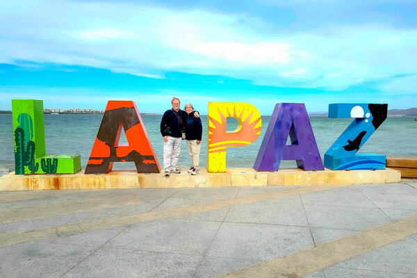 Старшая взрослая пара туристов, стоящая между буквами Ла-Пас, море на фоне голубого неба, зеленый, красный, желтый, фиолетовый цвета, наслаждаясь отдыхом, солнечный день в Ла-Пас, Нижняя Калифорния Sur Мексика