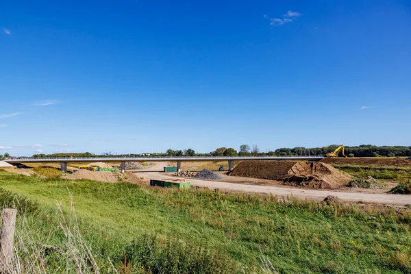 Landscape of rural road construction work area, a bridge, dirt road, crawler excavators and leafy trees against blue sky in background, sunny day in Dutch countryside in Meers, Elsloo, Netherlands