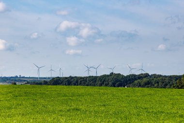 Puslu mavi gökyüzü, Belçika tarım arazisi Caestert Platosu, Riemst, Belçika 'da güneşli bir yaz günü, arka planda yapraklı ağaçlar ve yel değirmenleriyle kaplı yeşil bir ova. Van Groene enerjisi kavramı