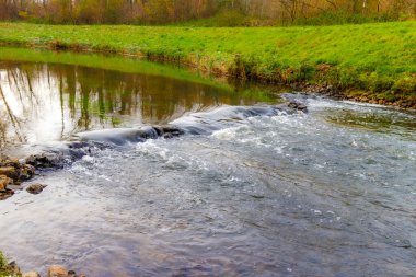 Gelenbeek deresindeki taşların üzerinden akan su, arka plandaki yeşil çimenler ve güz yaprakları, Hollanda doğa rezervi Geleenbeekdal, su yüzeyindeki yansıma, Schinnen, Güney Limburg, Hollanda