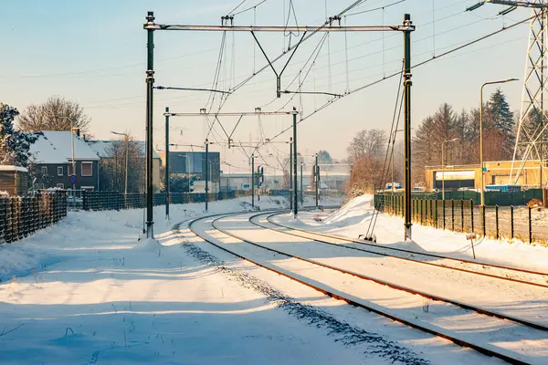 Electric cables on snow covered train tracks between houses and buildings, blurred misty background, sunny day after heavy snowfall in Beek - Elsloo, South Limburg, Netherlands