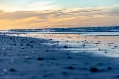 Sahil manzarasında gün batımı alttaki perspektiften, kumsaldaki kumlar bulanık ön planda, deniz dalgalı, ufuk ve arka planda altın turuncu bulutlar, Baja California Sur, Meksika