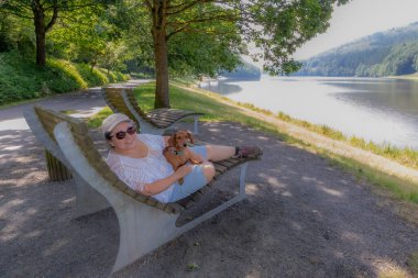 Ara veren turist, büyük bankın üzerinde dachshund 'unun yanında yatıyor, Stausee Bitburg gölünün kıyısında, kamera, patika ve arka planda yapraklı ağaçlara bakıyor, Almanya' da güneşli bir bahar günü