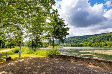 Park alanındaki dinlenme yeri, Echternach Gölü boyunca gezinti, beton zemin ve çitler, arka planda yapraklı ağaçlarla kaplı tepe, Lüksemburg 'da güneşli bir yaz günü.