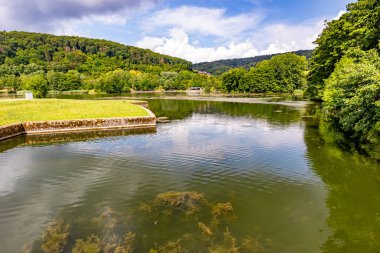 Parktaki küçük eğlence yarımadasını çevreleyen Echternach gölü, su yüzeyindeki algler ve yansımalar, arka planda yeşil yapraklı dağlar, Lüksemburg 'da güneşli bir yaz günü