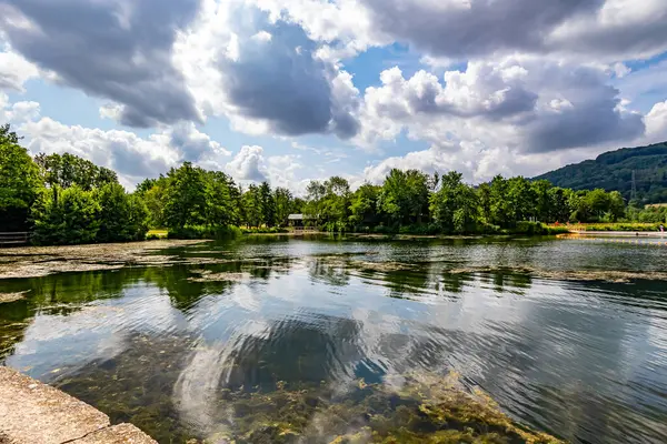 Echternach Gölü 'nün panoramik manzarasında su yüzeyinde yansımalar, sakin sularda algler, arka planda bol yapraklı ağaçlarla dolu tepeler, Lüksemburg' da bol bulutlu güneşli bir bahar günü.