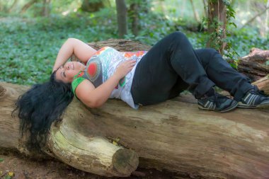 Tombul orta yaşlı bir kadın ağaç gövdesinde yatıyor. Arka planda yeşil bitki örtüsü var. Fotoğraf çekimi, uzun siyah dalgalı saçlar, renkli beyaz bluz ve siyah pantolon.