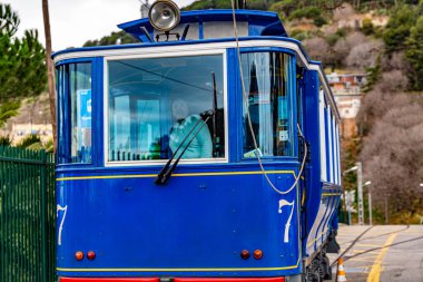 Yedi numaralı mavi tramvay bir tepeye doğru gidiyor. İçerideki bayan turist kameraya bakıyor. Arka planı bulanık olan dağlar, Katalonya Barselona 'da bir gün.