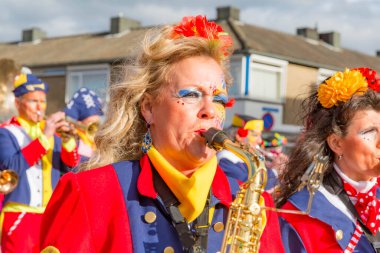 Beek, Güney Limburg, Hollanda. 3 Mart 2014. Yetişkin bir kadın, şehir sokaklarındaki karnaval geçidinde müzik grubunda flüt çalıyor. Mavi, kırmızı ve sarı renkli resmi elbiseler, bulanık arkaplan.