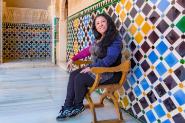 Orta yaşlı Latin Amerikalı kadın koridordaki ahşap sandalyede oturmuş sarayda mozaik duvarları olan kameraya, koyu renkli kıyafetlere Alhambra 'da turist, Granada, İspanya' da sakin bir gün.