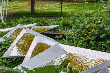Çağdaş metal çeşmesi, bitki örtüsü arasında akan su, arka planda yeşil çimen ve patika, Rheinpark halk parkı, Almanya 'nın Köln kentindeki bahar günü