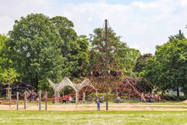 Köln, Almanya. 19 Mayıs 2024. Rheinpark 'taki çocuk parkındaki halat tırmanma oyunları, şehrin yeşil bölgesinde güneşli bir günün keyfini çıkaran aileler, arka planda mavi gökyüzüne karşı yapraklı ağaçlar.