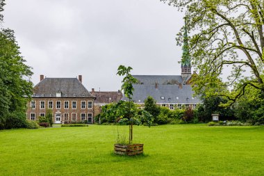 Val-Dieu Abbey bahçelerinin panoramik manzarası, ortasında küçük bir ağaçla kaplı yeşil çimenler, yapraklı ağaçlar, tapınak, arka planda kule ve bina, Belçika, Aubel 'de bulutlu bir gün.