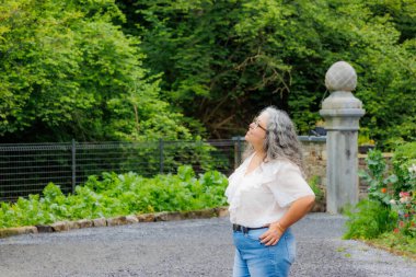 Üst düzey yetişkin kadın Val-Dieu Abbey bahçesinin girişine bakıyor, kot ve beyaz bluzlu günlük kıyafetler, bulanık arka planda yapraklı ağaçlar, Belçika, Aubel 'de bulutlu bir gün.