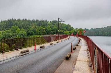Robertville Baraj Tepesi, bir kır yolu ve göl boyunca çitler arasında yaya geçidi, sisli arka planda yeşil yapraklı ağaçlar, Belçika, Waimes 'te bulutlu bir gün.
