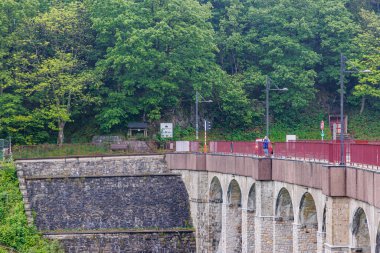 Robertville Barajı 'nın devasa kemerli duvarı, tepesinde araçlar ve yayalar için kırsal yol, Waimes, Belçika' da bulanık ve sisli arka planda yeşil yapraklı ağaçlar, bulutlu bir gün.